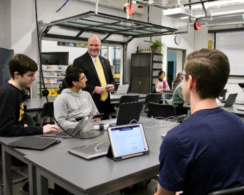 Three students seated at a table sharing a computer science project with Dr. Hunt, who is standing