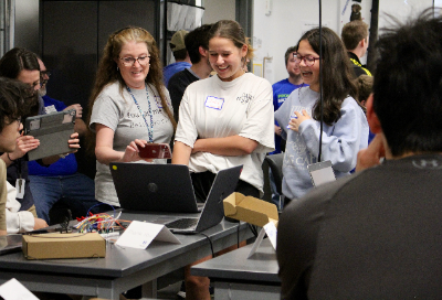 Two students smiling in front of a laptop with a teacher alongside them, holding up her phone to the screen to record a video, as others in the room look on