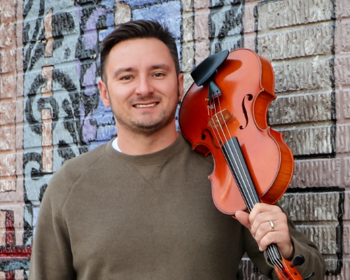 Chris Lape posing against a painted brick wall with a viola slung over his shoulder