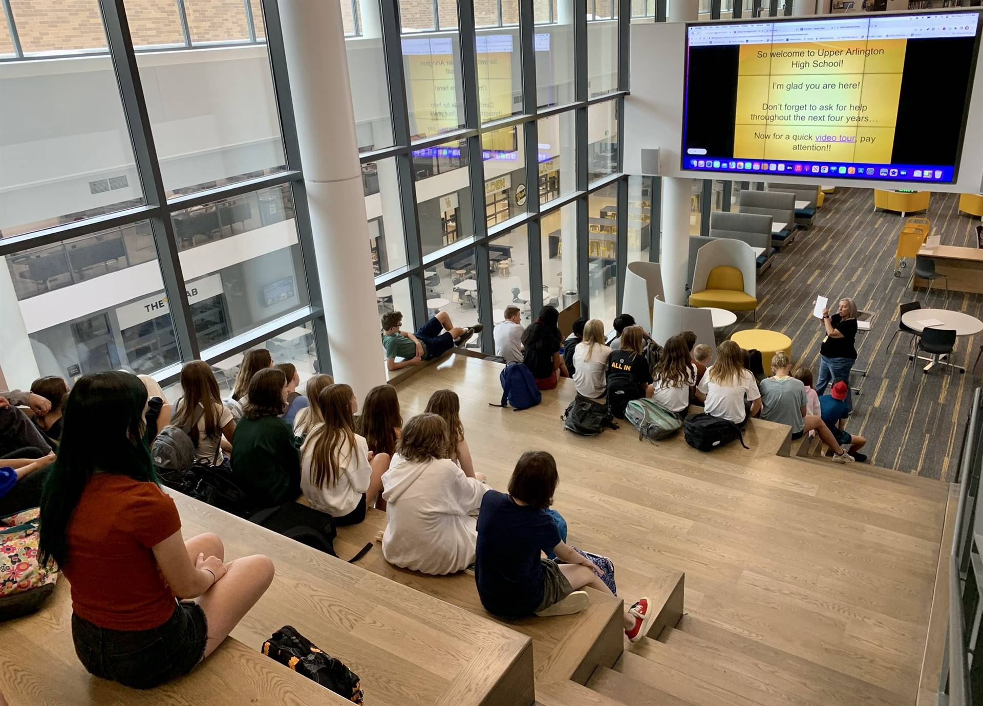 A photo of students sitting on large wooden stairs looking down on a presenter with a large screen o