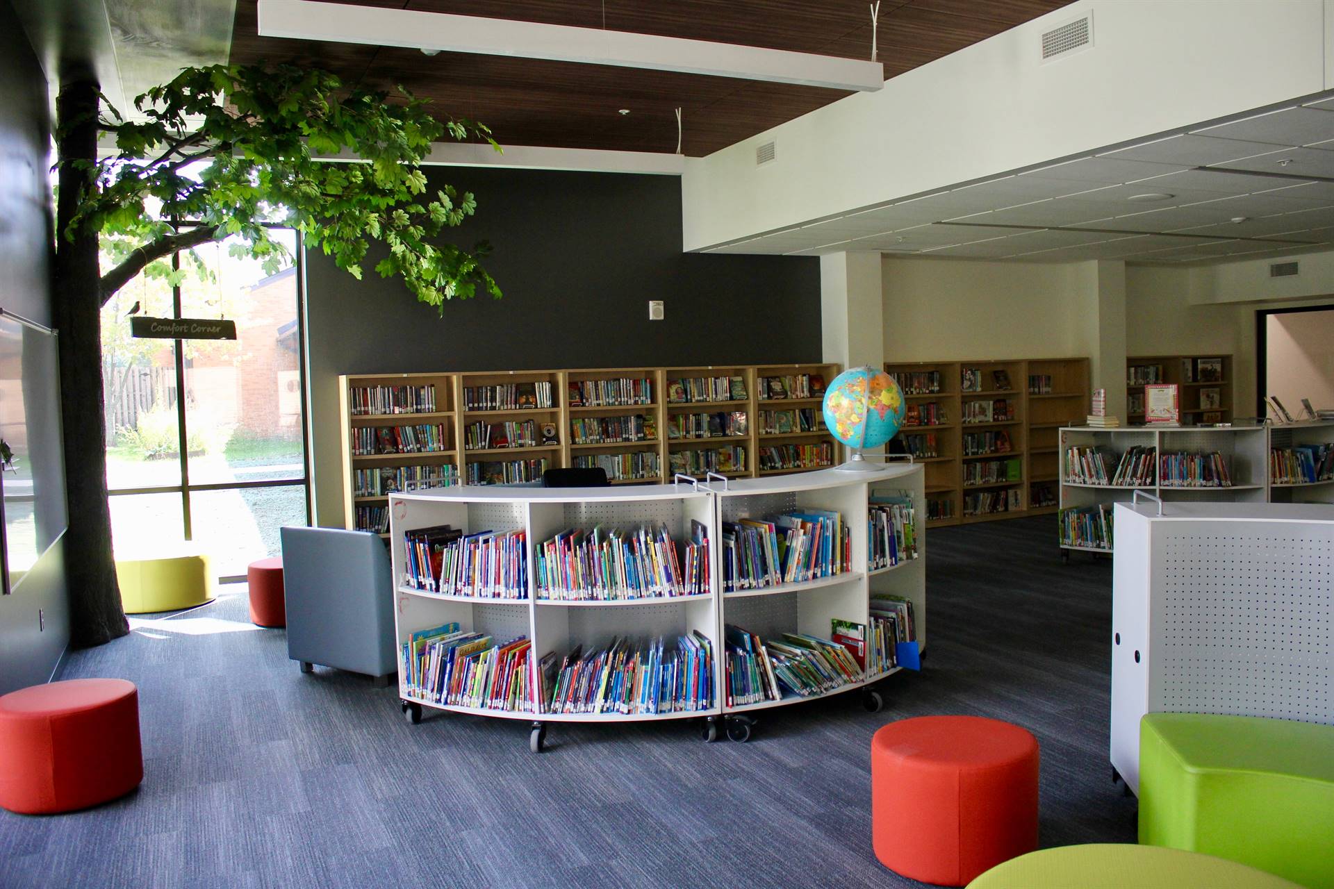 A photo of shelves of books, small colorful stools and a tree sculpture inside the media center
